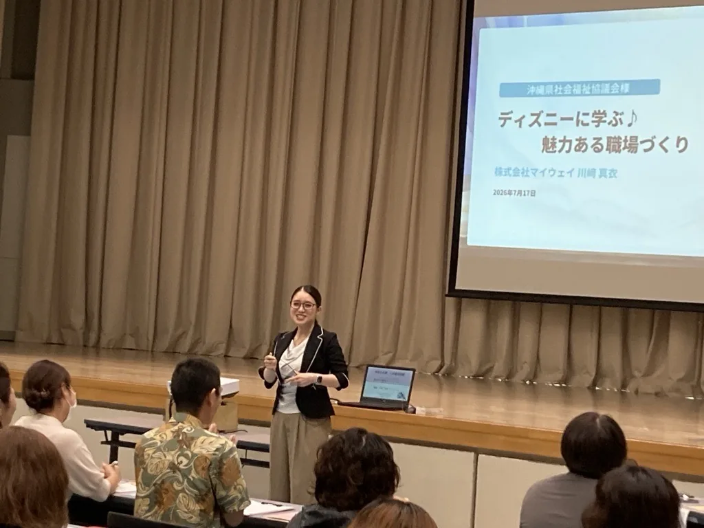 壇上で話す川崎真衣。スクリーンには「沖縄県社会福祉協議会様 ディズニーに学ぶ♪ 魅力ある職場づくり 株式会社マイウェイ 川﨑真衣 2026年7月17日」と映っている
